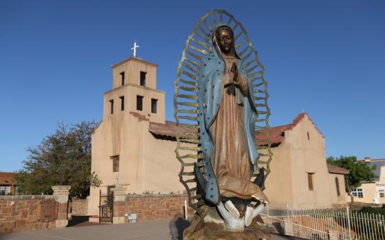 Statue of Our Lady of Guadalupe shown in front of large pueblo Church.