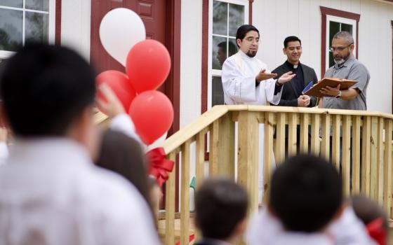 De Leon and others stand on the wooden porch of a prefab building saying a blessing; balloons and audience members frame shot.