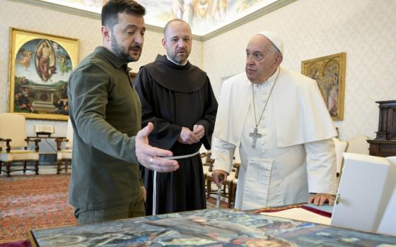 Francis, Zelenskyy, and a monk bend over a desk in the apostolic library to regard Zelenskyy's gift of an oil painting.