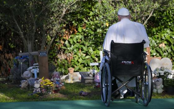 Francis pictured from behind, seated in wheelchair, alone and facing small graves adorned with votives.