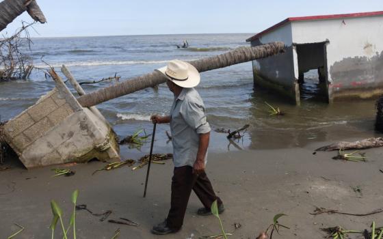 Man with walking stick foregrounded, in background are building ruins and the shoreline.