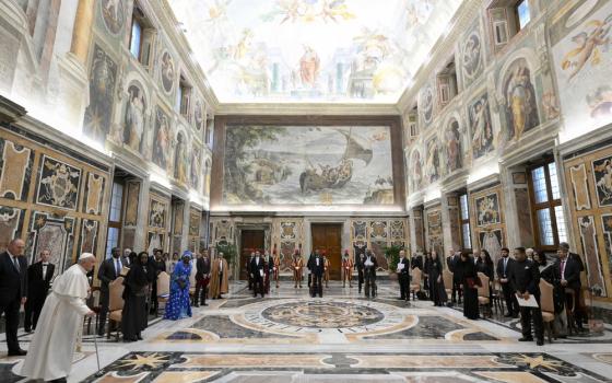 Large frescoed room; diplomats sit in u-shaped formation of chairs, Pope enters walking on cane. 