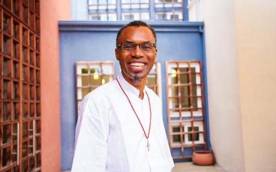 The priest wears white clothing and faces camera smiling.