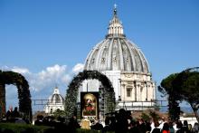 Leafy green arches are visible in front of the dome of St. Peter's Basilica with people sitting in the garden in front of an image of Mary