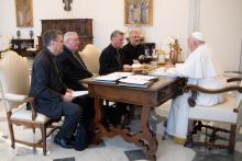 Pope Francis sits at his desk opposite four light-skinned men wearing black clerical garb