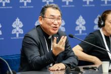 A smiling East Asian man wearing glasses and a clerical shirt and jacket speaks into a microphone