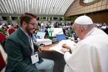 A young man with brown hair wearing a suit smiles at Pope Francis as he signs a document