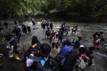 People wearing backpacks and other burdens walk through running water in jungle terrain. 