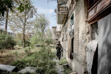 A man wearing protective gear walks along side an overgrown and crumbling building.