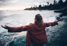 A woman stands with her arms outstretched, facing a coast in this photo illustration. (Unsplash/Nathan McBride)