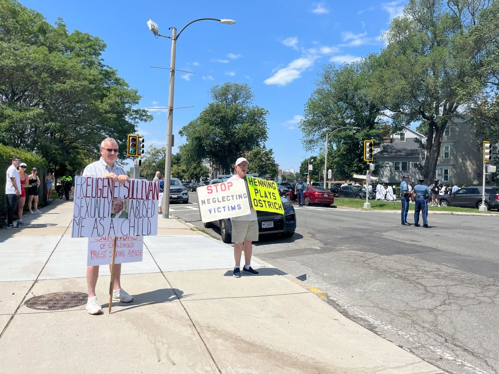 2 people hold protest signs referring to clergy sex abuse.