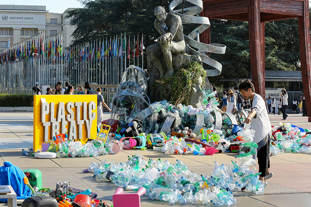 Benjamin Von Wong, right, a Canadian artist and activist, heaps piles of plastic waste onto a large sculpture that he designed in front of the United Nations office in Geneva on Aug. 11, 2025, during negotiations on a global treaty to address plastic pollution. (AP/Jennifer McDermott)