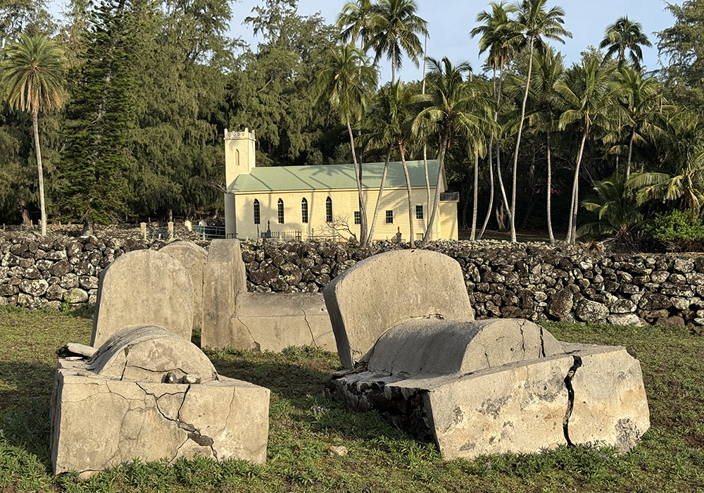 Graves are pictured at the Moku Puakala cemetery in Kalawao. In the background is the St. Philomena Catholic Church, St. Damien de Veuster’s main church which he helped to construct and expand, and the burial site of St. Damien. (National Park Service/Dewitt Jones)