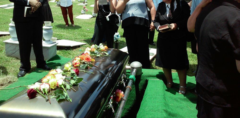 People stand around black coffin that has row of flowers on top.
