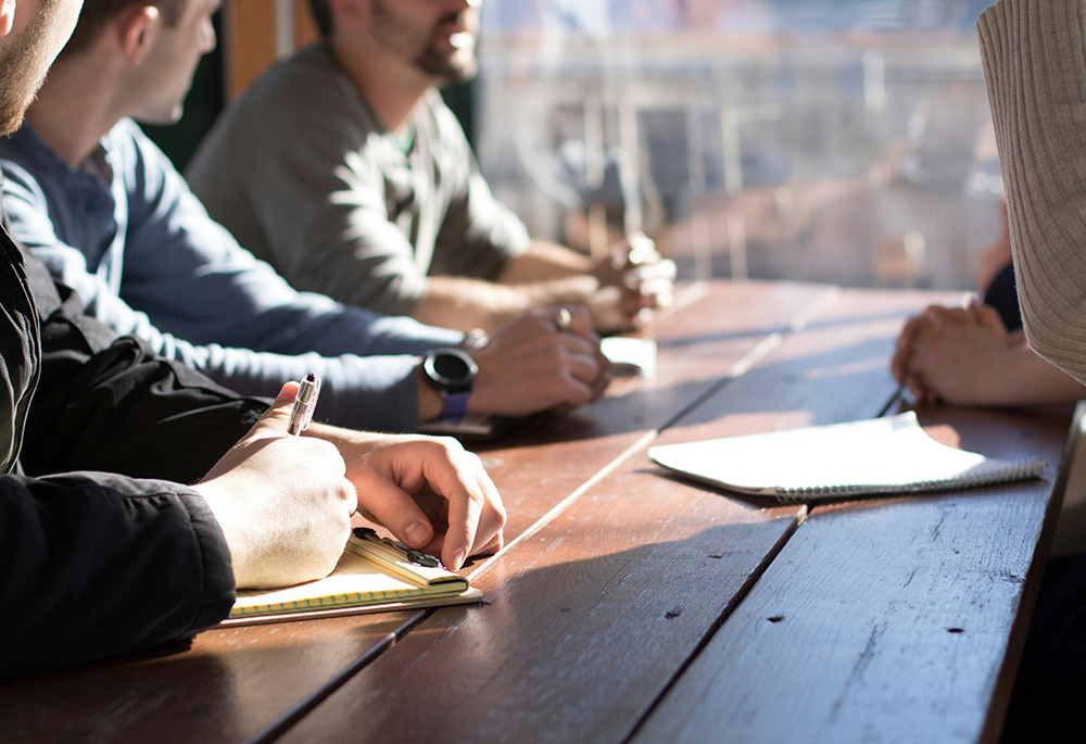 People sit with pen and paper in discussion at a table. (Unsplash/Dylan Gillis)