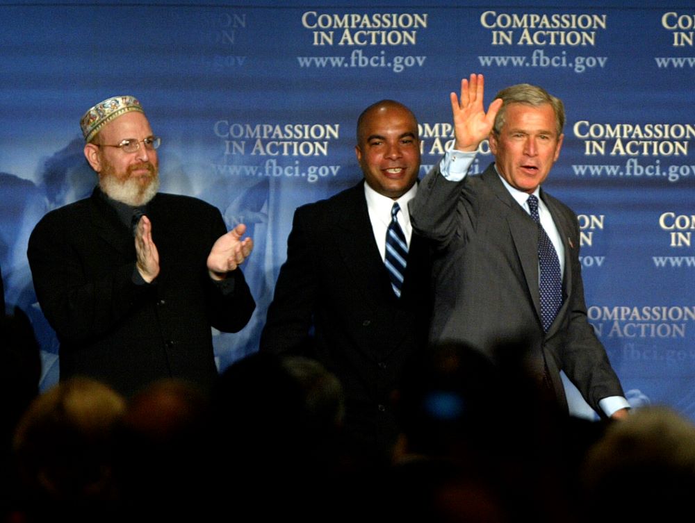 Rabbi Mark Borovitz, from left, and Julio Medina look on as President George W. Bush takes the stage to make remarks at the first meeting of the White House National Conference on Faith-Based and Community Initiatives, June 1, 2004, in Washington. (RNS/AP/Lawrence Jackson)