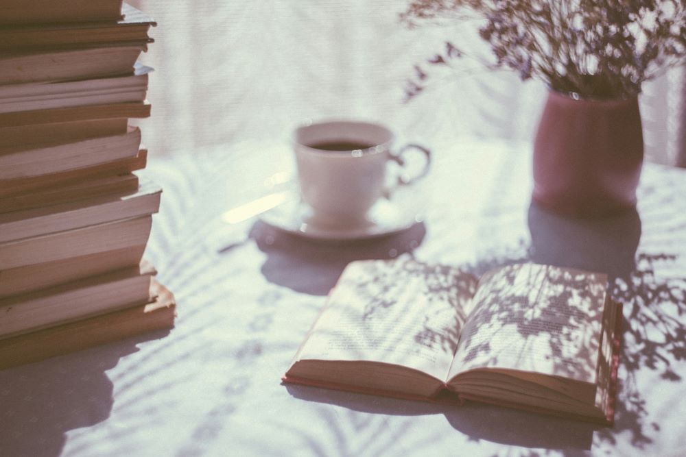 A pile of books, an open book, a cup of coffee or tea, and a plant sit on a table near a window.