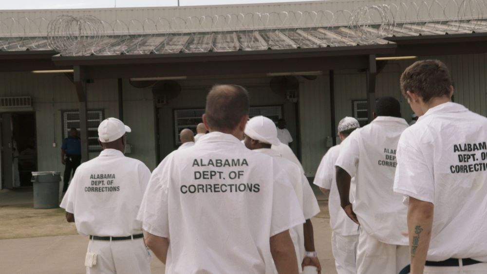 A still image shows a scene from "The Alabama Solution," nominated for a Best Documentary Feature Oscar, shows a group of men in white shirts that say, "Alabama Dept of Corrections" 
