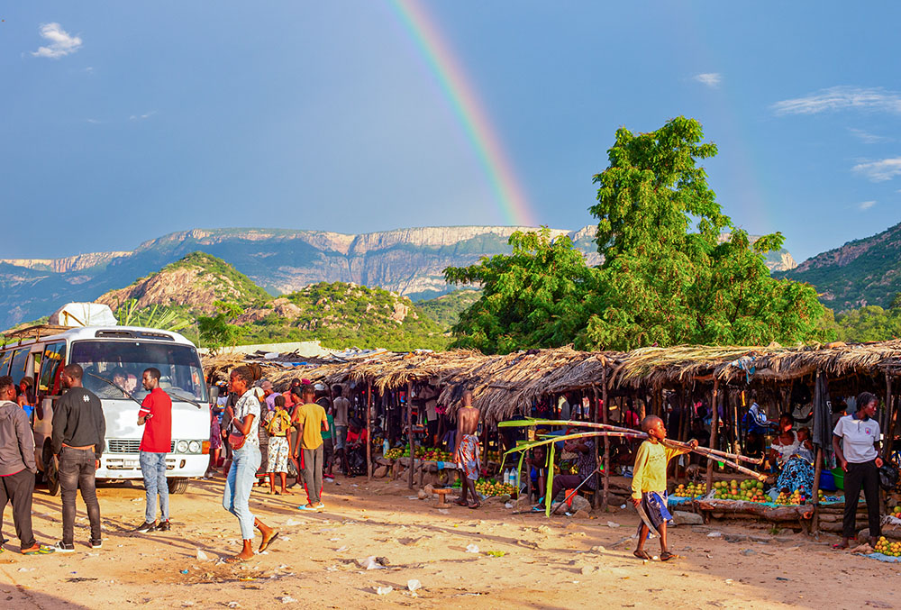 A rainbow hangs above mountains in the distance as people shop at a food market in Lubango, Angola, March 21, 2025. (Dreamstime/Joyfull)