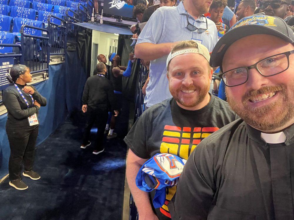 Fr. Danny Grover, right, with his brother Matthew before the May 18, 2026, Thunder game at which he delivered the invocation. Thunder star Shai Gilgeous-Alexander signs autographs in the background. (RNS/Courtesy of Danny Grover)
