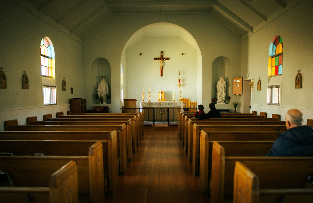 An older man sits in the back of a row of pews, facing the front of a nearly empty church. (Unsplash/Martin Jernberg)