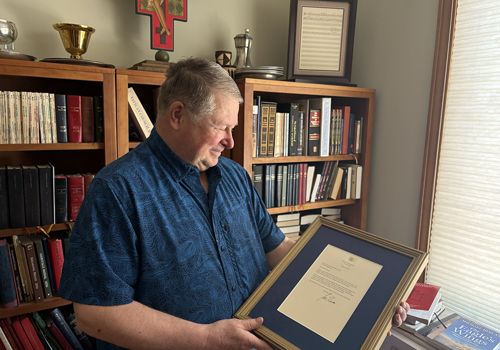 Author, composer and professor Fr. Michael Joncas holding a June 2024 letter from former U.S. president Joe Biden by the bookcase where he framed and displayed the original 1976 score of "On Eagle's Wings," in his apartment in St. Paul, Minnesota, May 2026. (NCR photo/Camillo Barone)