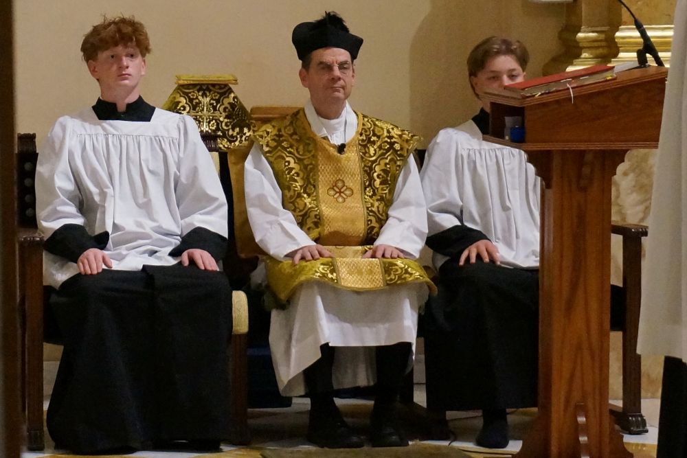 Fr. John Ubel, pastor of the Church of St. Agnes, wears the traditional biretta hat and vestments as he sits between altar servers during one of the Catholic parish's Latin Masses in St. Paul, Minn., June 28, 2026. (AP/Giovanna Dell'Orto)