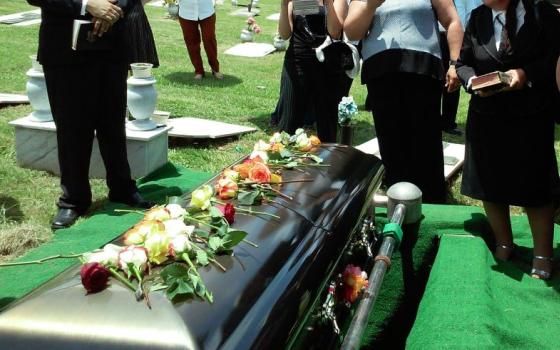 People stand around black coffin that has row of flowers on top.